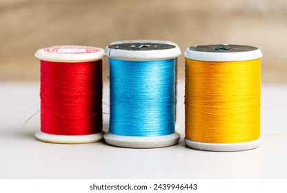 Three spools of thread in vibrant red, blue, and yellow stand in a row against a neutral background, illustrating the basic tools of sewing and textile arts.