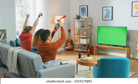 Three Sports Fans Sitting On A Couch In The Living Room Watch Green Chroma Key Screen TV Important Match, Using Horns, Having Fun And Cheering For Their Team.