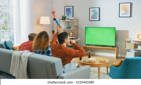 Three Sports Fans Sitting On A Couch In The Living Room Watch Green Chroma Key Screen TV Important Match, Using Horns, Having Fun And Cheering For Their Team.