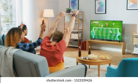 Three Sports Fans Sitting On A Couch In The Living Room Watch Important Soccer Match On TV, Cheering For Their Team, Celebrates Doing YES Gesture After The GOAL Brings Victory To His Team.