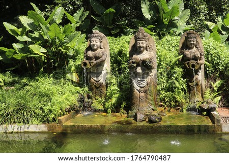 Three Statues in Bali Indonesia of a fountain with jungle in the background. Balinese statuette of a girl with a pot from which water flows. Pool decoration
