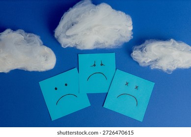 Three sticky notes with different expressions of sadness and depression overlapping under cotton clouds on a blue background.