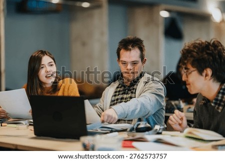 Three students work together at a table with a laptop and study materials. A collaborative study session shows young adults discussing documents and using a computer.