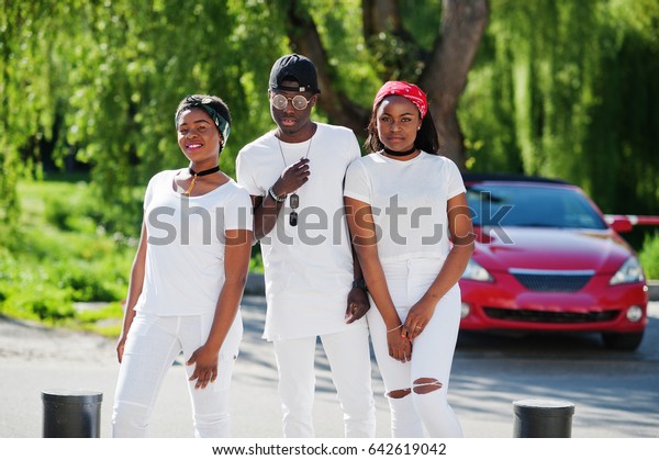 Three stylish african\
american friends, wear on white clothes against luxury red sport\
car. Street fashion of young black people. Black man with two\
african girls. 