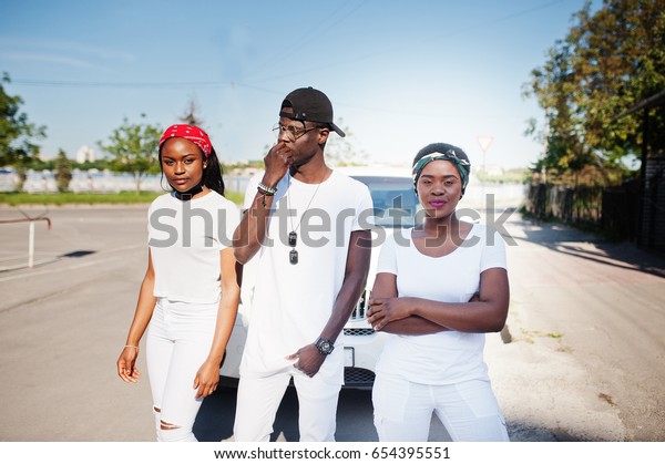 Three stylish african american\
friends, wear on white clothes against two luxury cars. Street\
fashion of young black people. Black man with two african\
girls.