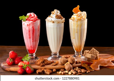 Three sweet milkshakes with nuts, caramel, strawberry and whipped cream at a wooden board on table background.