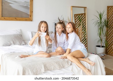 three teenage girls in white shirts and denim shorts having fun lying on bed and using phone for selfie in a stylish modern bedroom decorated minimalistic cozy Scandinavian design, gadget addiction