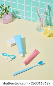 Three toothbrushes, floss picks, and colored toothpaste tubes laid flat against yellow surface with a green mosaic tile wall. Fun and cheerful hygiene photo.