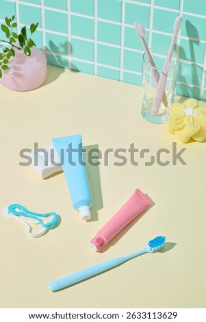 Three toothbrushes, floss picks, and colored toothpaste tubes laid flat against yellow surface with a green mosaic tile wall. Fun and cheerful hygiene photo.