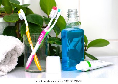 Three Toothbrushes In A Glass Cup Mouthwash A Tube Of Toothpaste And A Rolled Up Towel On The Dressing Table Against A Backdrop Of Green Foliage. High Quality Photo