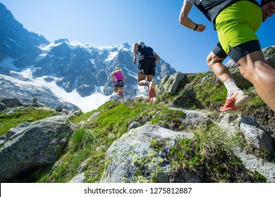 Three trail runners, two men and a woman, sprinting up a trail towards a huge mountain under the bright blue sky of summer in the Alps - Shutterstock ID 1275812887