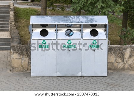 Three trash cans, in the shape of square boxes with three holes for garbage collection and labeled Glass, Plastic and Paper