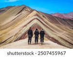 Three travelers admire the stunning Rainbow Mountain in Peru, with its vibrant layers of minerals and striking geological formations, under a clear blue sky.