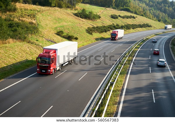 Three trucks and\
three cars driving on the highway beneath a hillside meadow and\
forest. View from above.