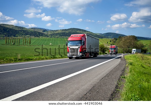 Three trucks driving on\
asphalt road between green fields in the countryside. Wooded\
mountains in the background. Sunny day with blue skies and white\
clouds.
