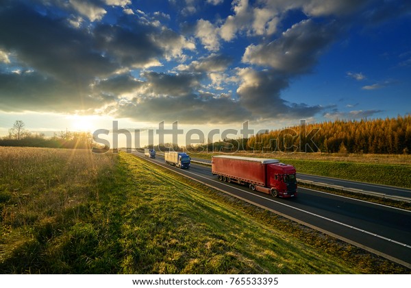 Three trucks driving on a highway in\
autumn landscape at sunset with dramatic\
clouds