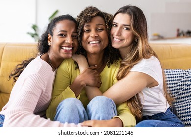 Three United Beautiful Smiling Women Sitting Together On Couch - Portrait Of Young Multiracial Girl Friends Hugging Each Other And Relaxing At Home - Friendship Concept - Focus On Faces