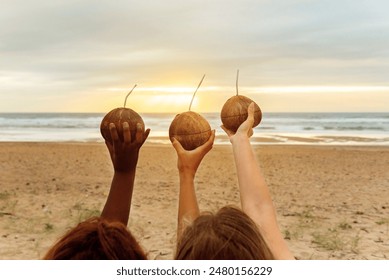 three unrecognizable young women of different races and skin tones holding in their raised hand a drink served in a coconut during a beach party at sunset. - Powered by Shutterstock