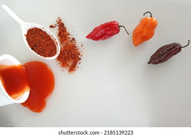 Three Varieties Of Hot Peppers Scotch Bonnet, Naga Morich, Bhut Jolokiaand Their Derivatives - Sauce And Ground Hot Pepper On A White Glass Surface