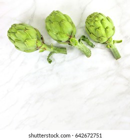 Three vibrant artichokes on a white marble texture with copy space, square photo