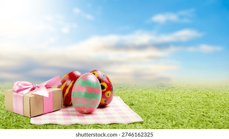 Three vibrant hand-painted Easter eggs and a gift box with a pink ribbon sit on a checkered picnic cloth in a lush green meadow under a bright blue sky. A beautiful scene of spring celebration
