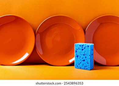 Three vibrant orange plates with contrasting blue sponge against an orange background, creating bold color composition. Perfect for cleaning and kitchen hygiene themes - Powered by Shutterstock