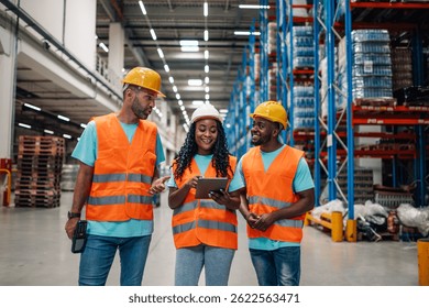 Three warehouse workers wearing safety vests and hardhats are walking through a large distribution center, using a digital tablet to discuss logistics and inventory management