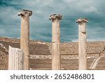 Three weathered ionic columns rising from ruins of ancient greek theater with stone steps under dramatic cloudy sky