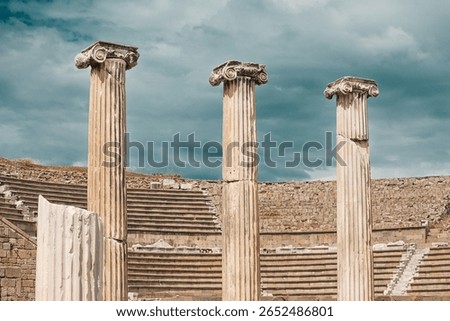 Three weathered ionic columns rising from ruins of ancient greek theater with stone steps under dramatic cloudy sky