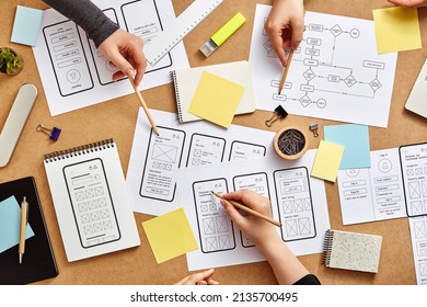 Three Web UX Designers Working Together At A Busy Desk With Numerous Mobile App Wireframe Sketches And User Flow. Top View.