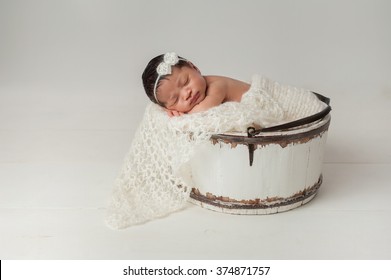 A three week old newborn baby girl sleeping in a little, wooden bucket. She is wearing a cream colored bow headband. Shot in the studio on a white background.