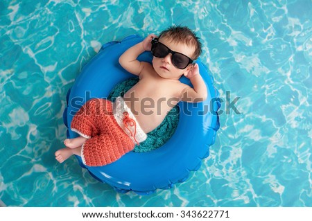 Three week old newborn baby boy sleeping on a tiny inflatable swim ring. He is wearing crocheted board shorts and black sunglasses.