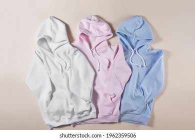 Three white, blue and pink pastel blank cotton hoodie laid out together isolated over pale beige background. Minimalism concept. Mockup. Horizontal shot