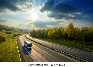 Three white buses traveling on the asphalt highway between deciduous forest in autumn colors under the radiant sun and dramatic clouds. View from above.                               