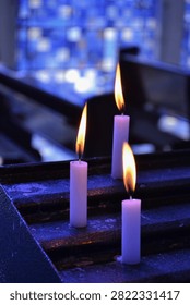 Three white candles burning inside a Catholic church with blue stained glass windows in the background, creating a spiritual and peaceful atmosphere.