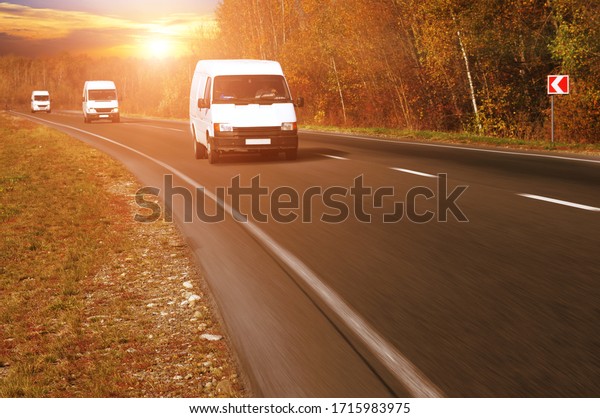 Three white commercial\
vans on a countryside road in motion with a forest against a night\
sky with a sunset