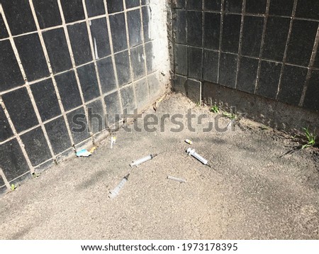 Three white plastic syringes with open needles lie on the gray textured asphalt in the corner near the house. The walls of the house are lined with black rectangular tiles. Drugs. Danger. Addiction. 