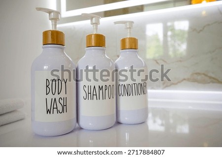 Three white refillable pump bottles labeled body wash, shampoo and conditioner with gold dispensers on bathroom countertop