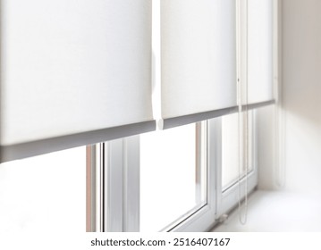 Three white roller blinds are pulled down over a window. The blinds are made of a thin, white fabric and have a gray bottom edge. The window is made of white vinyl and has a white trim.