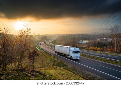 Three white truck driving on the highway winding through forested landscape in autumn colors at sunset