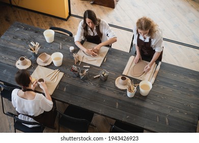Three women in aprons sitting by table, kneading clay and making earthenware at lesson - Shutterstock ID 1943426923