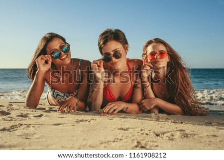Three women in bikini sunbathing lying on front at the beach holding their sunglasses. Woman on vacation relaxing at the beach wearing sunglasses having fun with sea in the background.