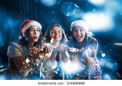 Three Women Blowing Confetti From Their Hands Indoors At Holiday Time.