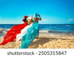 Three Women in Chiffon Colorful Dress flying on Wind on Beach. Ladies in Summer Hats looking at Sea. Girlfriend on Tropic Vacation. Bridesmaids in Silk Gown Back view