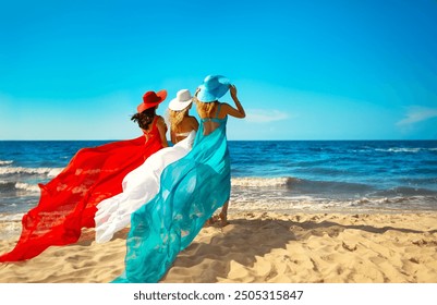 Three Women in Chiffon Colorful Dress flying on Wind on Beach. Ladies in Summer Hats looking at Sea. Girlfriend on Tropic Vacation. Bridesmaids in Silk Gown Back view - Powered by Shutterstock