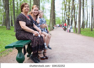 Three Women Different Ages Are Sitting On Bench In Park. Grandmother, Mother And Small Daughter