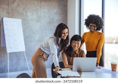 Three women of different ethnicities smiling and working together at a laptop in a bright, stylish office. - Powered by Shutterstock