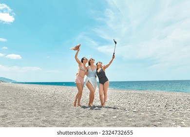 Three women friends walking on the beach, taking selfie and laughing on a summer day. Cheerful females enjoying together summer vacation near the sea.