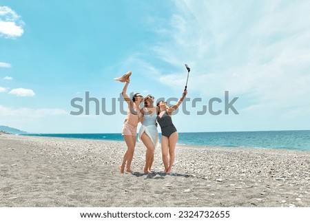 Three women friends walking on the beach, taking selfie and laughing on a summer day. Cheerful females enjoying together summer vacation near the sea.