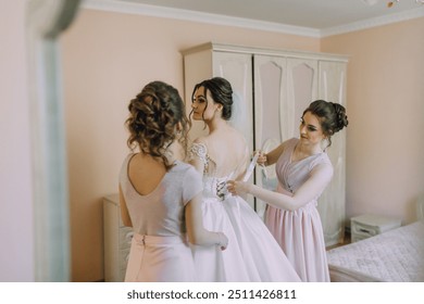 Three women are getting ready for a wedding. One of them is adjusting the bride's dress. The other two women are standing behind her, helping her. Scene is one of support and camaraderie - Powered by Shutterstock